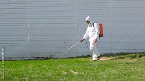 Person spraying pesticide on lawn field wearing a disposable protective suit. Treatment of grass from weeds and insects. Pest control. Insecticide sprayer with a proper protection. Garden care season.