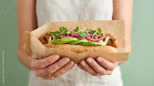 A woman is holding an elegant plant-based vegan hot dog wrapped in kraft paper. A top-down view of this healthy fast food item topped with avocado, fresh herbs, and pickled onions. AI generated.

