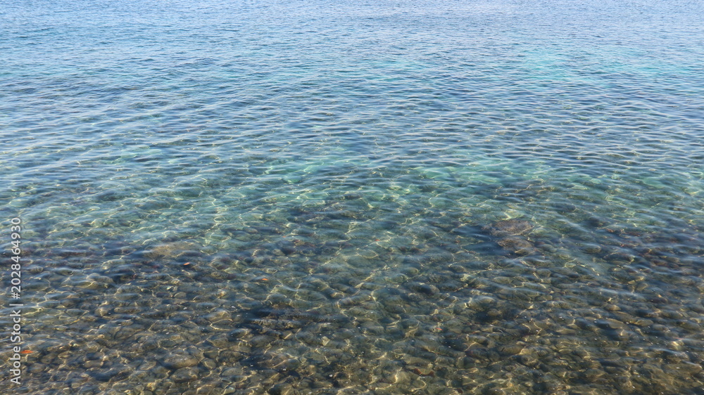 Obraz premium Clear water gently ripples over rocks below. Shallow sea reveals a bed of smooth stones. Light dances across the water's surface. Ideal for tranquil nature or travel visuals. No people, no animals