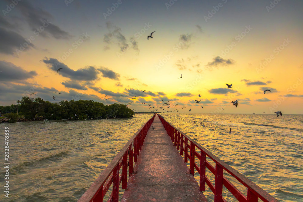 Fototapeta premium Red bridge to the sea with seagulls at sunset,A scenic view of a long red wooden bridge stretching into the sea during a dramatic sunset, with a flock of seagulls flying in the golden sky.