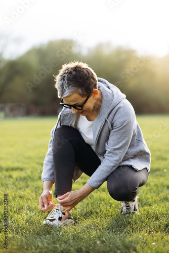 Active senior woman crouching in a park, adjusting her running shoe and preparing for outdoor fitness