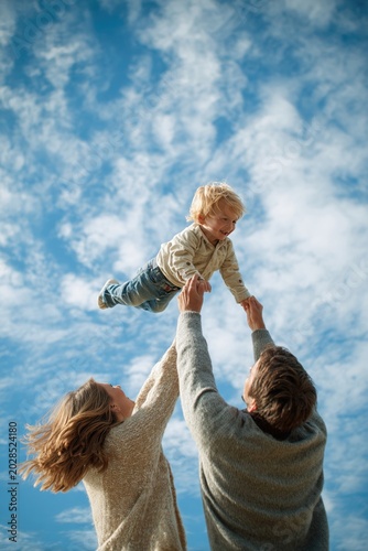 Parents lifting toddler into air in park, bright blue sky, action shot, crisp detail, happy carefree moment