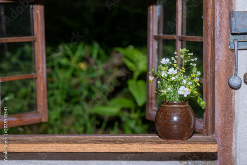 A small ceramic vase with tiny white wildflowers sits on a rustic wooden window sill looking out into a green garden.
