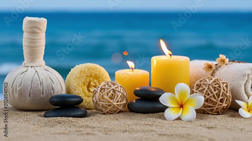 Spa treatment items arranged on sandy beach near ocean. Zen stone, burning candle, herbal compress and frangipani flower for wellness. Relaxation therapy concept.