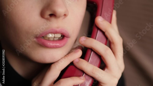 Extreme close-up of a boy's face talking on a red vintage telephone. The focus is on the lips and hands of the child, who is firmly holding the retro receiver during a call.