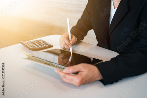 Close-up of hands using a stylus pen to e-sign a document, financial contract, or drawing on a digital tablet screen, with a blurred calculator on the wooden desk. Paperless and fintech concept.