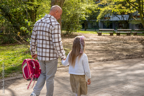 Grandfather walking hand in hand with granddaughter across sunny park, little girl wearing a pink school backpack, Walking to school spring day, education concept, family bonding, outdoor lifestyle