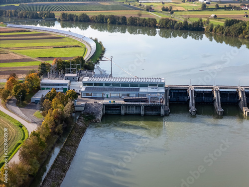 Aerial view captures hydroelectric power plant by a river surrounded by fields and trees. Views show roads and agricultural land nearby.