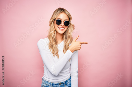 young woman in sunglasses pointing upward on pink background. Fashion, summer lifestyle and positive advertising concept with copy space.