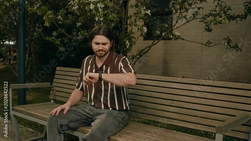Couple sits on bench in park while man gestures angrily after long wait. Tense date scene shows disappointment and conflict.