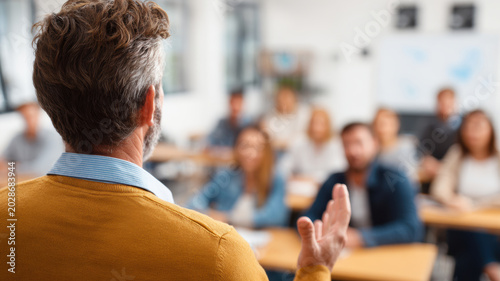 Education training class. Knowledge learning improvement study. A man in a yellow sweater standing in front of a classroom full of people seated at desks, attentively listening to a presentation.