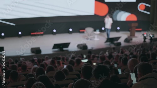 Audience watching keynote presentation at busy tech conference, speaker on stage, large screen visuals, attendees using phones and laptops, dim auditorium lighting.