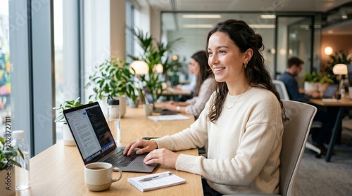A woman sits smiling at a desk with a laptop in a modern office environment surrounded by plants and colleagues.