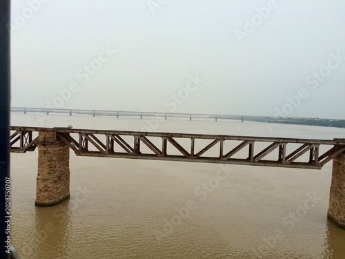 Vászonkép Railway bridge with metal truss and stone pillars spanning a wide river, captured from a moving train, with calm water and hazy sky in the background