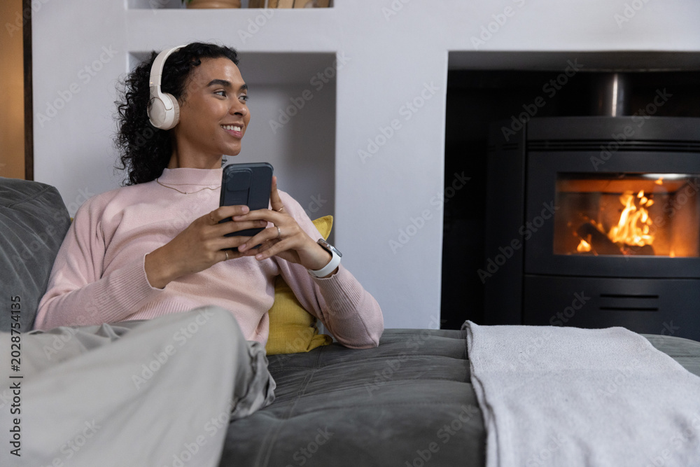 Fototapeta premium Nonbinary adult reclining on gray sofa at home wearing headset holding dark phone near fireplace