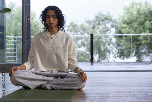 Non-binary adult meditating on green mat near glass doors wearing cream hoodie and gray sweatpants