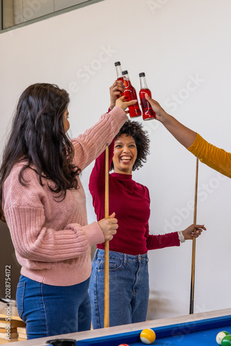 Diverse friends in casual clothes raising bottles and holding cues by pool table in recreation room