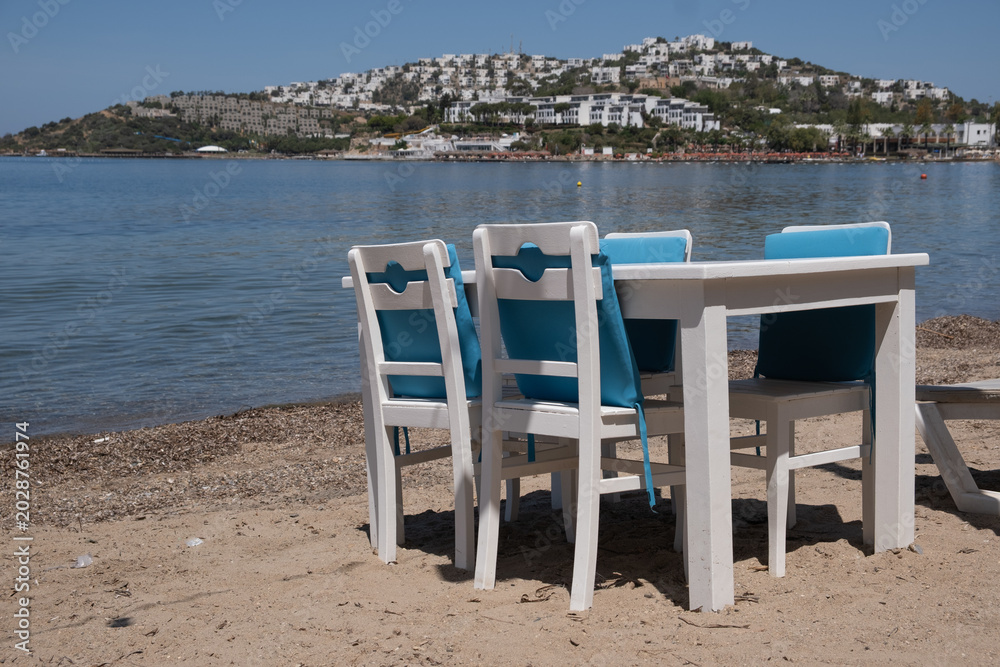 Fototapeta premium Table and chairs on sandy beach near sea with background of white buildings on hill