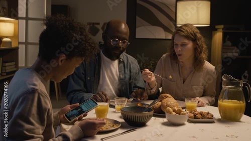 Side view shot of Black teenage boy using smartphone at dinner table and showing funny video to smiling parents sharing interests, copy space