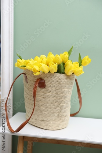 Yellow tulips in wicker bag on white table near green wall