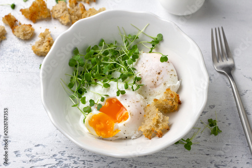 Poached eggs with microgreens and bread croutons in white bowl on light rustic background