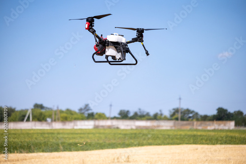 An unmanned agricultural drone flies over a cultivated field under a clear blue sky during daytime operations to spray crops with liquid fertilizer or pesticides from its onboard white tank