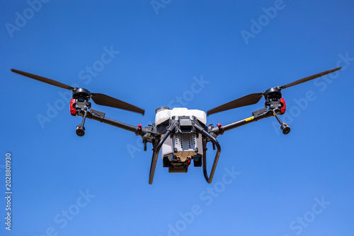 An unmanned aerial agricultural drone equipped with a white liquid chemical spraying tank flies through a clear blue sky while performing precision farming operations over regional crop fields