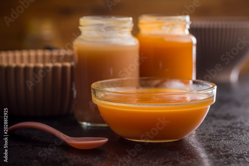 Creamy Pumpkin Soup In Glass Bowl With Jars Of Puree And Spoon On Dark Surface