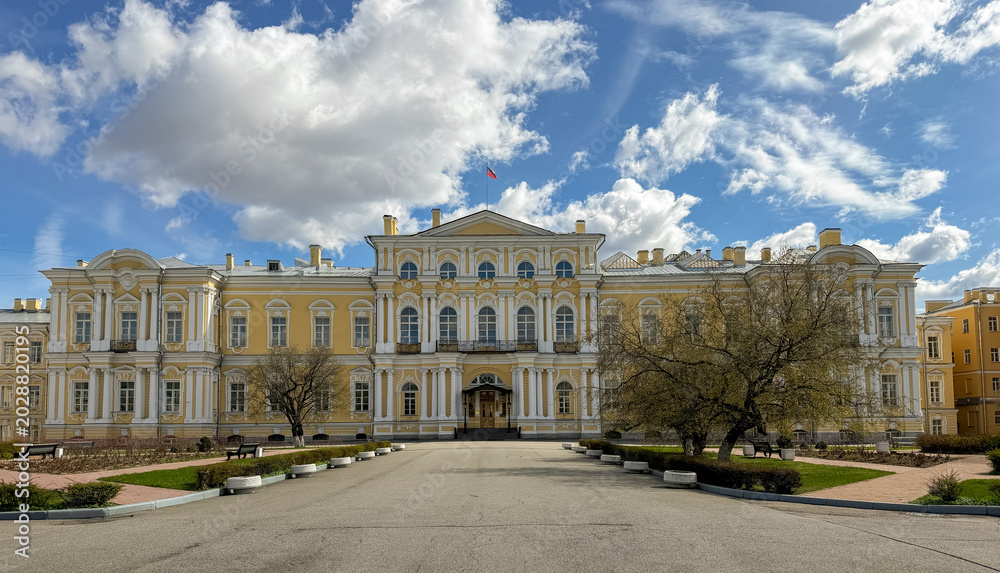 Naklejka premium Historic yellow building features classical architecture and large windows, surrounded by trees and a paved pathway under a blue sky with clouds