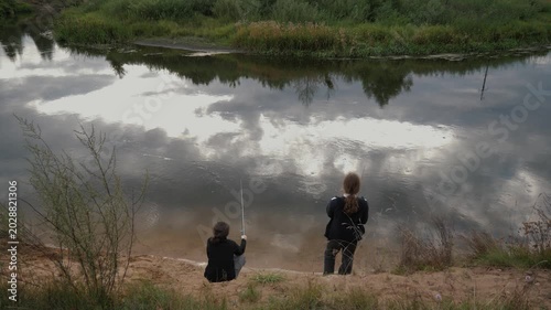 A fishing lesson for young explorers on a tranquil riverbank. Two teenagers fish in the calm river water under an overcast summer sky