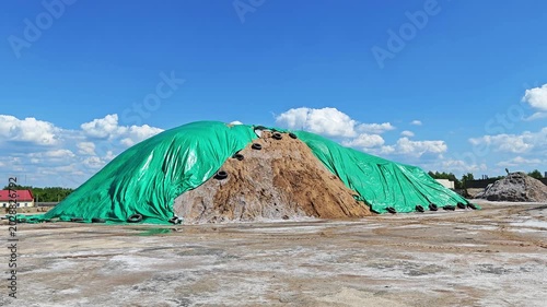 Huge sand-salt pile for winter road maintenance covered with green PVC film against rain, blowing in the wind under blue sky. 4K footage of seasonal deicing material storage.