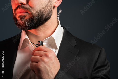 Photos Bearded security guard or intelligence agent pressing a lapel microphone on his collar, using a radio earpiece for communication