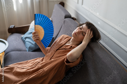 Tableau sur toile Stressed woman suffering from summer heat and migraine at home, lying on sofa waving paper fan for cooling, unwell female on couch with headache touching forehead and fanning herself with hand fan