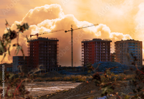 Obra urbana con grúas y edificios residenciales bajo un dramático cielo anaranjado y humeante. Imagen editorial sobre desarrollo urbano y construcción inmobiliaria para noticias sobre crecimiento