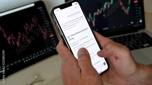 Man hands holding smartphone with AI chatbot showing trading analysis text in front of stock market monitors with charts. Concept of fintech, AI trading assistant, investment and market analysis.