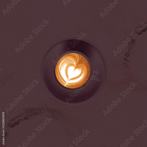 A top-down view of a latte with heart-shaped foam art sits in a dark mug on a textured surface. The warm glow of the coffee illuminates the creamy latte art.