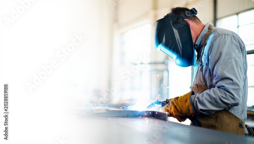 Konstfotografi Male welder in grey jacket and dark helmet leaning over a workbench welding a steel plate with blue arc and sparks in a brightly sunlit industrial factory with generous copy space