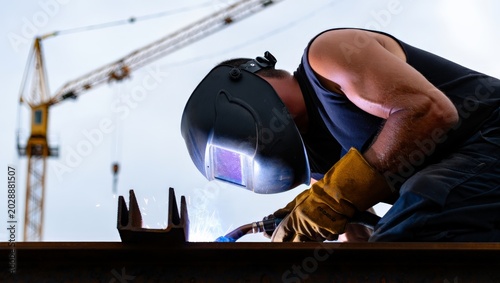 Obraz na plátně Low-angle shot of a muscular welder in a black auto-darkening helmet and leather gloves welding a steel beam on a construction site with a yellow tower crane rising against an overcast sky