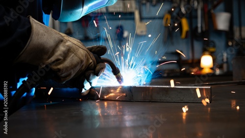 Konstfotografi Close-up of a welder in a blue-tinted helmet and worn leather gloves MIG welding a steel box section, vivid blue arc and orange sparks scattering across a dark workshop with tool wall behind