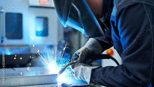 Quadro em tela Dynamic close-up of a welder in blue coverall and dark helmet MIG welding a steel plate with a vivid blue arc and sparks flying toward the camera in a blue-lit factory environment