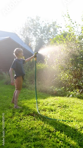 Vertical video of funny boy pouring cool water on himself and playing with garden hose with sprinkler in sunny backyard. Happy child having fun with water splashes. Summer outdoor activity for kids.