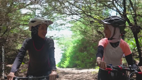 Two asian women cycling forest trail smiling and talking, helmeted, wearing sporty jerseys and hijabs, sidebyside ride