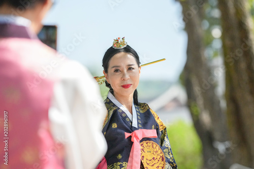 Woman in traditional Korean attire smiles gently. She wears ornate headdress and intricate robes. Her expression radiates warmth and grace. Soft sunlight enhances the natural outdoor setting