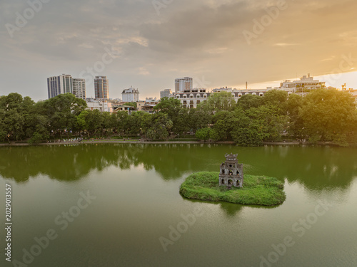 Golden evening sunlight illuminates Hanoi Hoan Kiem district with colonial buildings, modern towers, and tree lined streets beside the lake.