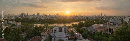 Wide panoramic sunset over Hanoi skyline and Hoan Kiem Lake district with glowing sky, dense urban development, and reflective water.