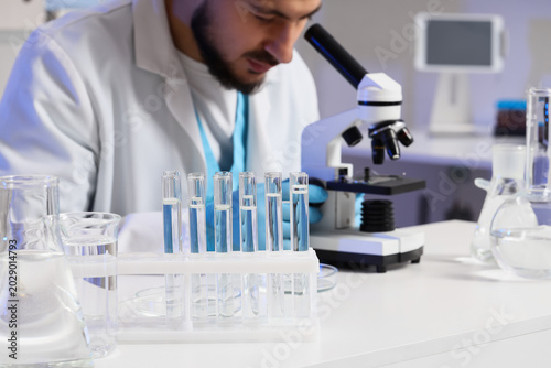 Male scientist with microscope and test tubes of water sample working at tabl...