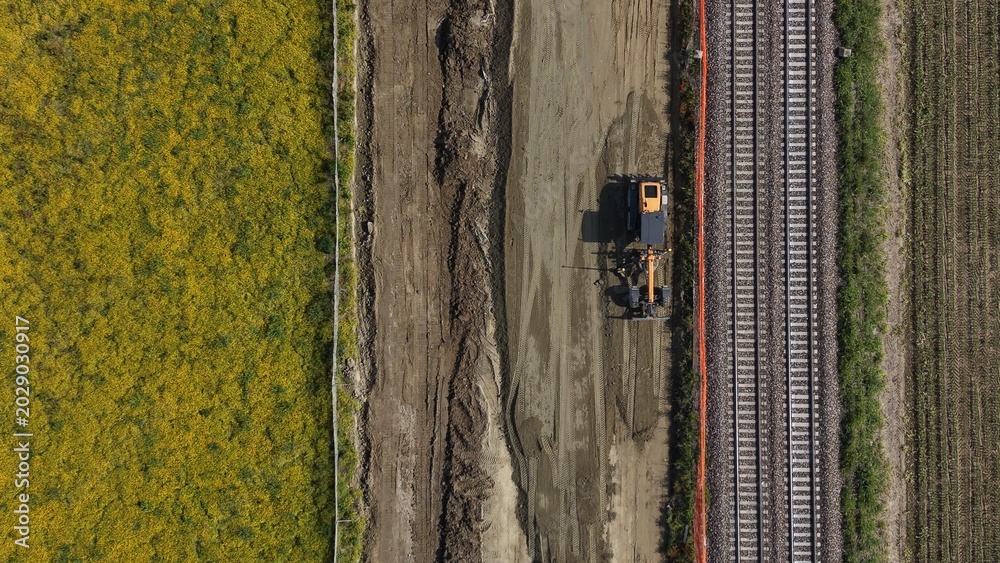 Fototapeta premium Railway Construction Aerial View: An aerial perspective reveals a construction site next to a railway track, where heavy machinery operates on bare earth adjacent to a field of yellow crops.