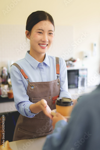 Asian female barista smiling serving takeaway coffee cup to customer at cafe counter wearing apron providing friendly service at coffee shop business