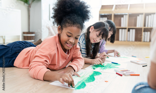 Group of happy, multicultural kids lying on floor and doing colorful drawings with crayons during creative indoor school activity.