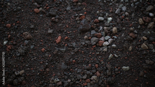 Fotografie Top-down texture view of dark volcanic soil mixed with small rocks, multi-colored pebbles, and red pumice pieces on a rough ground surface